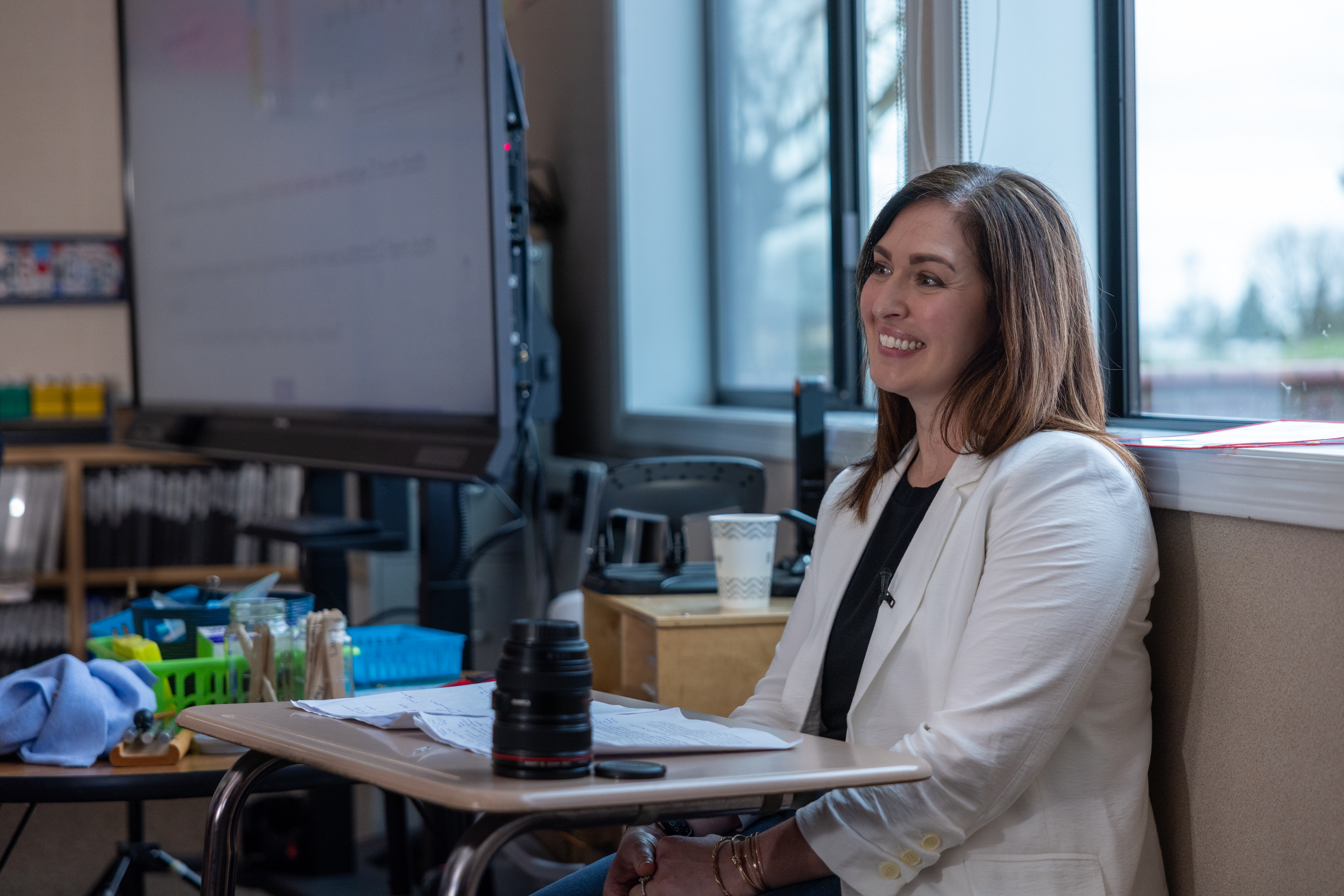 A woman in a white blazer sits at a desk in a classroom, smiling and looking to the side. A camera lens sits on the desk next to her, suggesting she may be involved in photography or videography. The classroom setting implies she could be a teacher or presenter, possibly giving a lecture or demonstration.